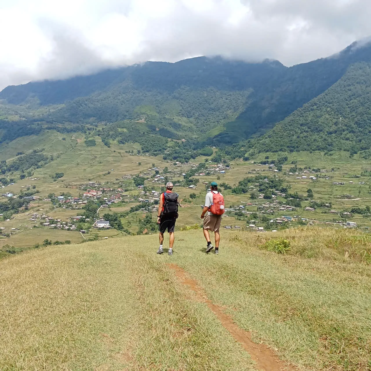 Two people with backpacks walking a grassy track above a wide valley of fields and houses
