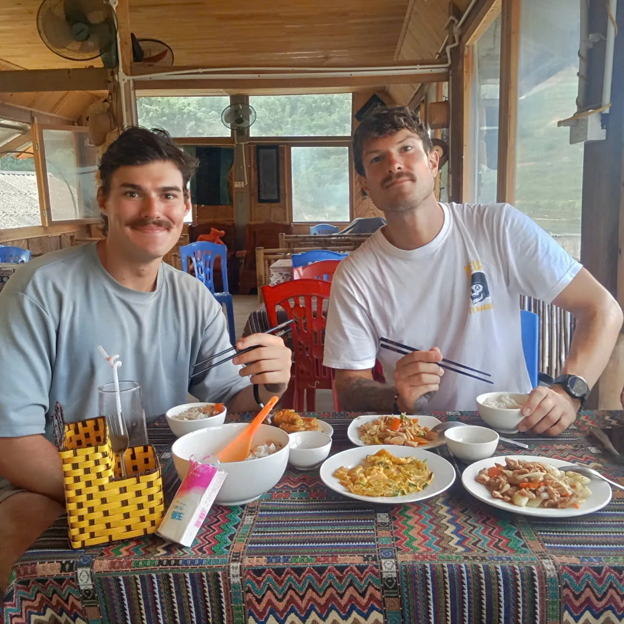 Two people eating rice and shared dishes at a wooden table in an open-sided room