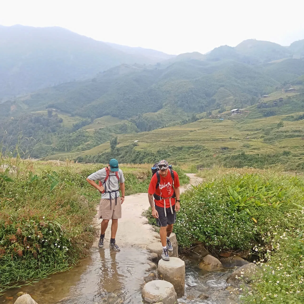 Two people with backpacks crossing a shallow stream on round concrete stepping stones
