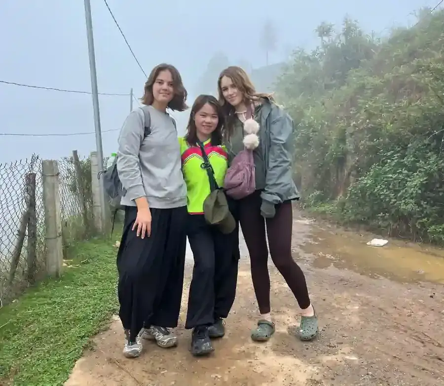 Three young travelers smiling together with their local Sapa guide on a misty mountain trail during a homestay trekking experience