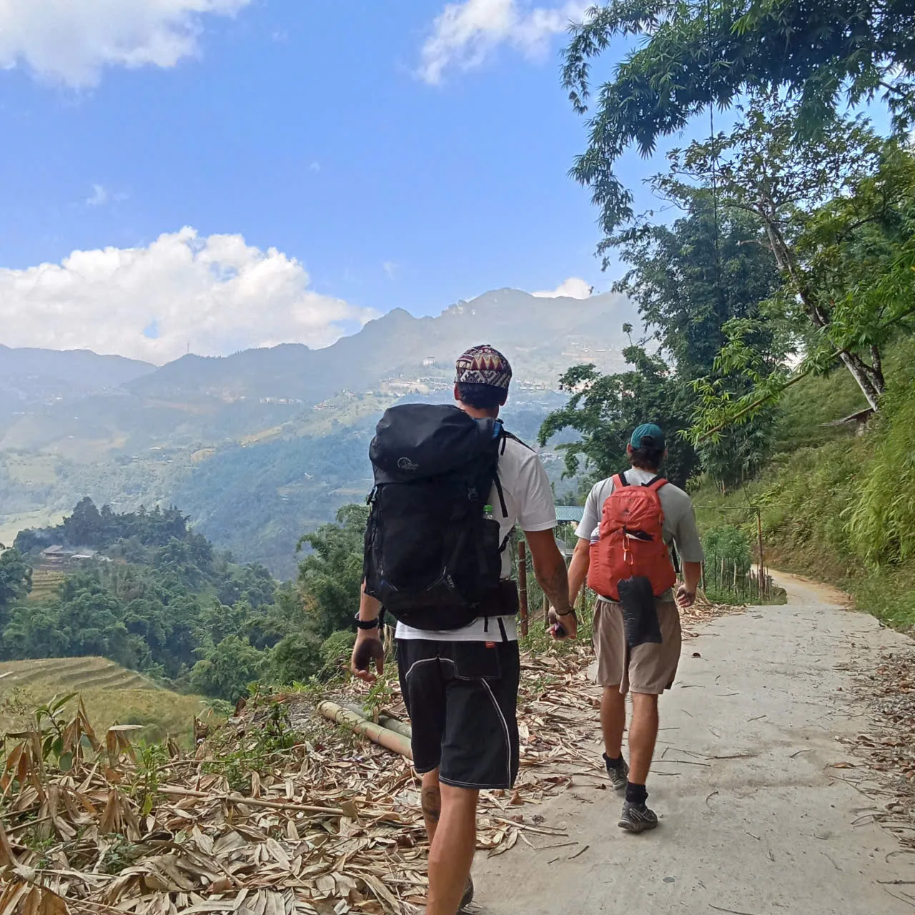 Two people with backpacks walking away along a concrete path above rice terraces and forested mountains